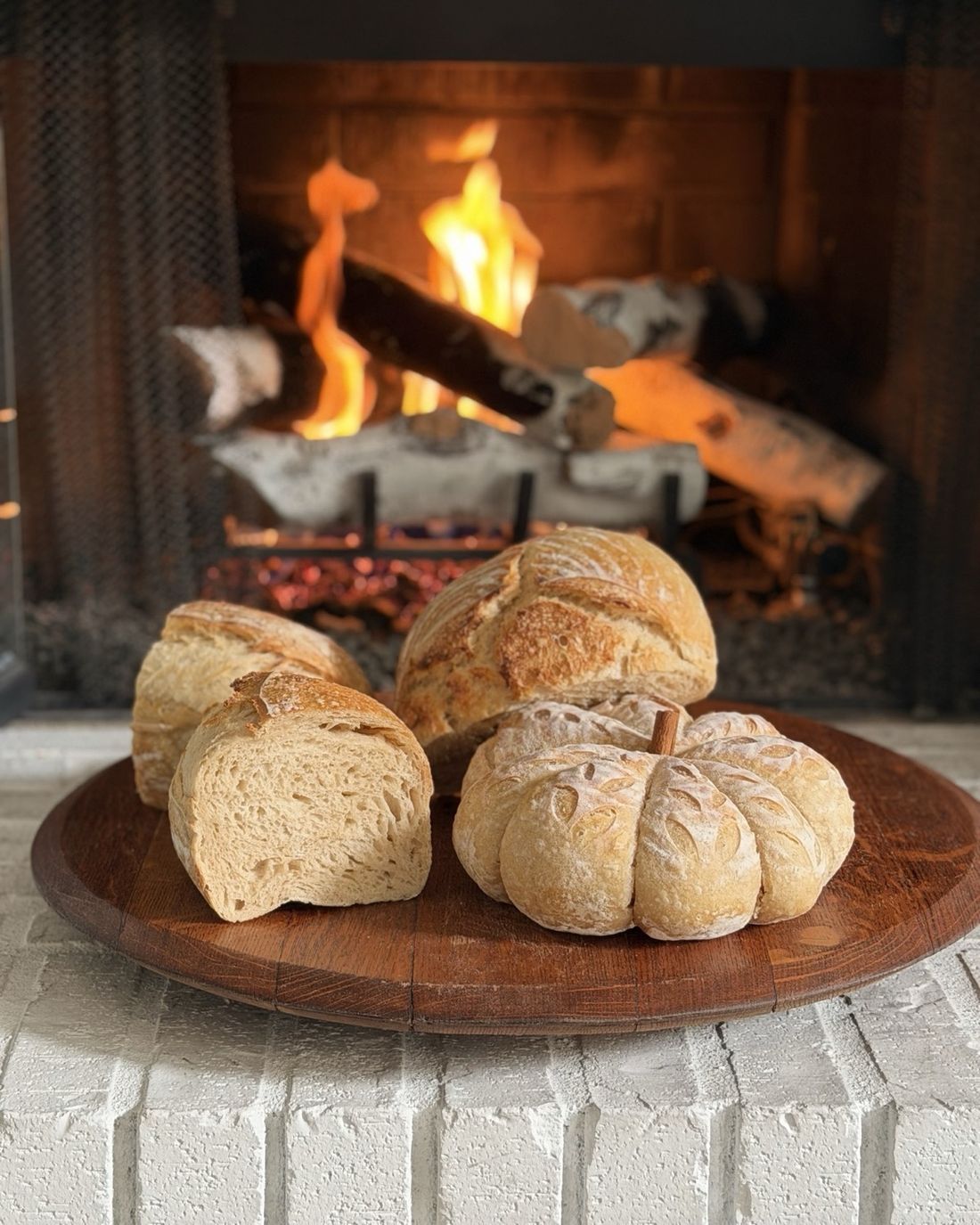 Sourdough loaves and a pumpkin-shaped loaf on a wooden board in front of a fireplace